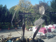 Kranichbrunnen: In einer Schale aus Metall liegen faustgroße Kieselsteine. Zwischen den Steinen steht ein Kranich aus Metall, der in die Luft schaut. Aus einem geraden Metallstab mit einer Spirale am oberen Ende läuft Wasser über den Kranich.