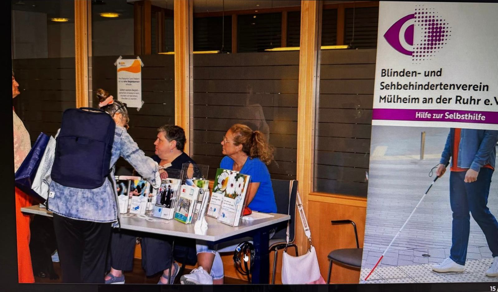 Ein Informationsstand des Blinden- und Sehbehindertenvereins Mülheim an der Ruhr e. V. ist aufgebaut. Hinter dem Tisch sitzen zwei Frauen: links Maria St. Mont (Vorsitzende) und rechts Nicole Schmitz-Siegfried (Vorstandsmitglied). Auf dem Tisch stehen zahlreiche Flyer, Informationsmaterialien und ein Kalender mit Bildern von Blindenführhunden. Eine Besucherin mit großem Rucksack und Einkaufstasche beugt sich über den Tisch und spricht mit den beiden. Rechts steht ein Roll-Up mit dem Logo des Vereins, dem Schriftzug „Hilfe zur Selbsthilfe“ und einem Bild eines Mannes mit Blindenstock.
