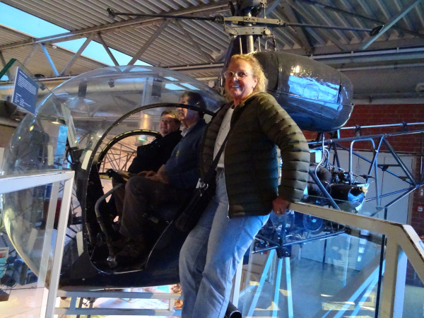 Ein älterer Hubschrauber im Ausstellungsraum, vermutlich ein Museumsstück. Drei Personen sind zu sehen: Zwei sitzen in der Kabine des Hubschraubers, eine Frau steht vorne an der Einstiegstreppe und lächelt. Die Kabine des Hubschraubers ist durch eine große, runde Glaskuppel deutlich sichtbar. Im Hintergrund sieht man moderne Gebäudestrukturen mit Metallträgern und Lampen. Die Atmosphäre wirkt freundlich und interessiert, vermutlich ein Museumsbesuch.
