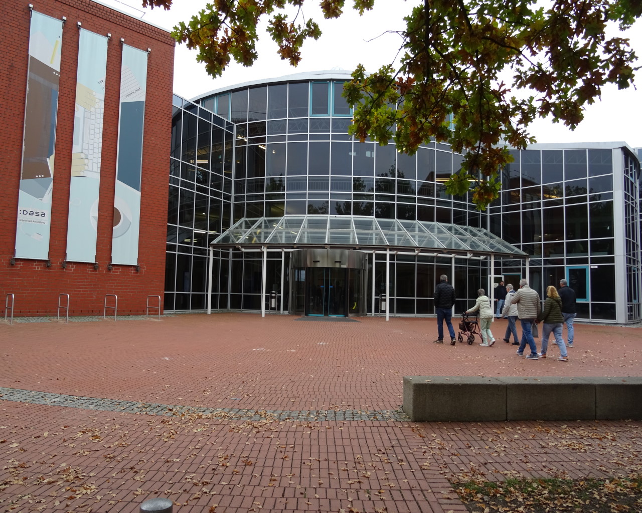 Eingangsbereich eines modernen Gebäudes mit viel Glasfassade und roten Klinkersteinen. Über dem Haupteingang ist ein Glasvordach angebracht. Links an der Wand hängen drei große vertikale Banner mit teilweise erkennbarem Text, darunter das Wort DASA. Vor dem Eingang gehen einige Menschen, darunter auch eine Person mit Rollator. Im Vordergrund liegt ein gepflasterter Platz mit ein paar verstreuten Herbstblättern und etwas Laub, ein Baum mit buntem Laub ragt von oben ins Bild.