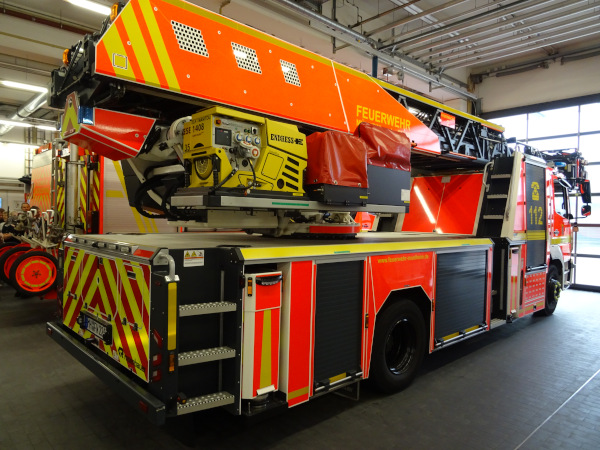 Großes Feuerwehr-Drehleiterfahrzeug in einer Fahrzeughalle. Das Auto ist in leuchtendem Rot mit gelben und weißen Warnstreifen lackiert und steht seitlich leicht schräg nach hinten versetzt. Auf der rechten Fahrzeugseite befinden sich mehrere große, schwarze Rollläden für Gerätekästen, eingefasst von gelben Kantenmarkierungen. Über den Gerätekästen verläuft ein gelber Streifen mit der Internetadresse „www.feuerwehr-muelheim.de“. Hinten am Fahrzeug ist eine große Klappe mit schrägen gelb-roten Warnstreifen und darunter ein deutsches Kfz-Kennzeichen angebracht. Rechts hinten ist eine senkrechte Reihe von Trittstufen aus Metall montiert. Am Heck und an der Fahrzeugseite sind zahlreiche Rück- und Blinklichter sowie Reflektoren verteilt. Auf dem Fahrzeugaufbau ist der Drehleiter-Aufbau montiert. Unter dem ausfahrbaren Leiterpaket befindet sich ein gelbes Aggregat mit vielen Knöpfen, Schaltern, Anzeigen und Piktogrammen; darauf steht unter anderem „ENDRESS ESE 1408“. Daneben sind graue und rote Technikmodule zu sehen, teilweise mit roten Abdeckhauben geschützt. Die eigentliche Leiter liegt im eingefahrenen Zustand auf dem Dach, mit mehreren schwarzen Gittersegmenten. Auf dem seitlichen Leiterkasten steht groß „FEUERWEHR“ in Gelb. An der rechten Führerhausseite ist die Notrufnummer „112“ mit einem gelben Telefonsymbol auf rotem Grund zu erkennen. Das Fahrzeug steht in einer hohen Halle mit offenem Tor im Hintergrund, mehreren Deckenlampen und Leitungen unter der Decke. Links am Bildrand sind weitere Feuerwehrgeräte mit roten Rädern in einer Reihe abgestellt. Großes Feuerwehr-Drehleiterfahrzeug in einer Fahrzeughalle. Das Auto ist in leuchtendem Rot mit gelben und weißen Warnstreifen lackiert und steht seitlich leicht schräg nach hinten versetzt. Auf der rechten Fahrzeugseite befinden sich mehrere große, schwarze Rollläden für Gerätekästen, eingefasst von gelben Kantenmarkierungen. Über den Gerätekästen verläuft ein gelber Streifen mit der Internetadresse „www.feuerwehr-muelheim.de“. Hinten am Fahrzeug ist eine große Klappe mit schrägen gelb-roten Warnstreifen und darunter ein deutsches Kfz-Kennzeichen angebracht. Rechts hinten ist eine senkrechte Reihe von Trittstufen aus Metall montiert. Am Heck und an der Fahrzeugseite sind zahlreiche Rück- und Blinklichter sowie Reflektoren verteilt. Auf dem Fahrzeugaufbau ist der Drehleiter-Aufbau montiert. Unter dem ausfahrbaren Leiterpaket befindet sich ein gelbes Aggregat mit vielen Knöpfen, Schaltern, Anzeigen und Piktogrammen; darauf steht unter anderem „ENDRESS ESE 1408“. Daneben sind graue und rote Technikmodule zu sehen, teilweise mit roten Abdeckhauben geschützt. Die eigentliche Leiter liegt im eingefahrenen Zustand auf dem Dach, mit mehreren schwarzen Gittersegmenten. Auf dem seitlichen Leiterkasten steht groß „FEUERWEHR“ in Gelb. An der rechten Führerhausseite ist die Notrufnummer „112“ mit einem gelben Telefonsymbol auf rotem Grund zu erkennen. Das Fahrzeug steht in einer hohen Halle mit offenem Tor im Hintergrund, mehreren Deckenlampen und Leitungen unter der Decke. Links am Bildrand sind weitere Feuerwehrgeräte mit roten Rädern in einer Reihe abgestellt.