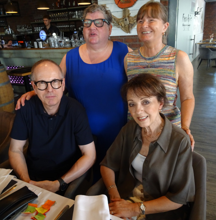 Vier Personen posieren gemeinsam für ein Gruppenfoto in einem Restaurant. Vorne sitzen von links Stefan Wagner in einem dunklen Poloshirt und Gaby Chauvet in einer olivgrünen Bluse. Hinter ihnen stehen Maria St. Mont in einem blauen Kleid und Tamara Ströter in einem gestreiften ärmellosen Oberteil. Alle blicken in die Kamera und lächeln. Im Hintergrund sind der Tresen des Restaurants sowie Regale mit Gläsern und Flaschen zu sehen.