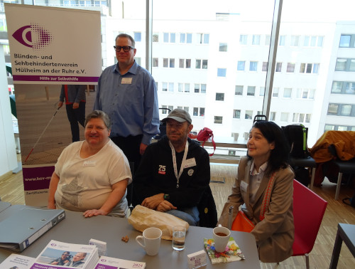 Mitglieder des BSV Mülheim Vorstandes, sowie der Vorsitzende des Landesverbandes BSV Nordrhein.  Vier Personen an einem Infostand eines Blinden- und Sehbehindertenvereins in einem hellen Raum.  Links sitzt eine ältere Frau mit heller Haut und kurzen grauen Haaren an einem Tisch. Sie trägt ein weißes T‑Shirt mit einem glitzernden Stern auf der Vorderseite und lächelt. An ihrer Brust ist ein Namensschild befestigt.  Hinter ihr steht ein Mann mittleren Alters mit heller Haut, kurzen Haaren und dunkler Brille. Er trägt ein hellblaues Hemd und ebenfalls ein Namensschild. Rechts von ihm ist ein Roll‑Banner aufgestellt. Darauf ist oben ein stilisiertes, lila‑weißes Auge mit Punkten zu sehen. Darunter steht: „Blinden- und Sehbehindertenverein Mülheim an der Ruhr e.V. Hilfe zur Selbsthilfe“. Unten ist eine Person mit Langstock beim Gehen zu erkennen sowie die Internetadresse „www.bsv-mh.de“ und das Wort „Zertifizierte …“ (der Rest ist verdeckt).  Vorne rechts am Tisch sitzt ein Mann mit heller Haut, Brille und grauer Schirmmütze. Er trägt eine dunkle Kapuzenjacke mit Aufdrucken und ein Namensschild an einem weißen Lanyard. Vor ihm liegt ein brauner Papiersack, daneben ein kleines Glas mit Wasser und eine weiße Tasse.  Ganz rechts sitzt eine Frau mit heller Haut und langen, dunklen, leicht gewellten Haaren. Sie trägt einen beigefarbenen Blazer über einem hellen Oberteil, ein Namensschild und eine orange Umhängetasche mit gemustertem Gurt.  Auf dem grauen Tisch im Vordergrund liegen mehrere Broschüren und Flyer des Vereins, ein Aktenordner, eine weiße Kaffeetasse, ein weiteres Glas und eine bunte Papierserviette.  Im Hintergrund befinden sich große Fenster, durch die mehrstöckige weiße Wohn- oder Bürogebäude mit vielen rechteckigen Fenstern erkennbar sind. Auf einer Bank vor den Fenstern liegen mehrere Rucksäcke und Jacken.