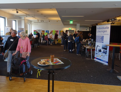 Messe- oder Infoveranstaltung in einem großen, hellen Raum. Im Vordergrund steht ein rundes, hohes Stehtischchen mit einem schwarzen Tisch