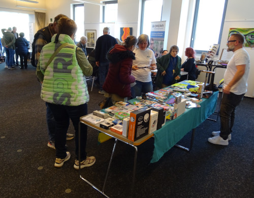 Ein Verkaufs- oder Infostand in einem Innenraum mit mehreren Menschen darum herum. Vorne steht ein rechteckiger Tisch mit einer türkisfarbenen Tischdecke. Darauf liegen viele kleine Produkte dicht nebeneinander, zum Beispiel Schachteln mit technischen Geräten, bunte verpackte Gegenstände, lange dunkle Stäbe oder Löffel, Schlüsselanhänger und verschiedene Hilfsmittel; einige größere Kartons stehen an der vorderen Tischkante. Links nah am Tisch steht eine Person mit hellgrüner, gesteppten Weste, auf der groß das Wort „SPIRIT“ senkrecht aufgedruckt ist, dazu dunkle Hose und helle Turnschuhe. An der rechten Tischseite steht ein Mann mit weißem T‑Shirt, dunkler Hose und weißen Turnschuhen; er trägt Bart und Brille und wirkt wie Standpersonal. Mehrere weitere Personen stehen auf der dem Betrachter abgewandten Längsseite des Tisches, teils in Winterjacken, teils in weißen T‑Shirts; eine Frau in roter Steppjacke betrachtet etwas in den Händen, eine andere in weißem T‑Shirt spricht mit ihr. Im Hintergrund befinden sich weitere Tische mit Menschen, die sich unterhalten oder etwas begutachten. An den Wänden hängen mehrere bunte Bilder oder Gemälde. Rechts hinter dem Haupttisch steht ein Roll‑Up‑Banner mit der Aufschrift „ORCAM“ und darunter kleineren Text, offenbar Werbung für ein Hilfsmittel für blinde oder sehbehinderte Menschen. Der Raum wirkt wie ein Veranstaltungs- oder Ausstellungsraum mit Teppichboden, großen Fenstern und Tageslicht.