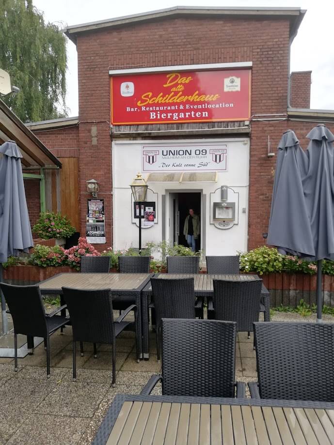 Eingang zu einem Backsteingebäude mit der Aufschrift „Das alte Schilderhaus – Bar, Restaurant & Eventlocation – Biergarten“. Vor dem Eingang stehen schwarze Gartentische und -stühle, teils mit geschlossenen Sonnenschirmen. Über der Tür hängt zusätzlich ein Schild mit „Union 09 Mülheim an der Ruhr“.