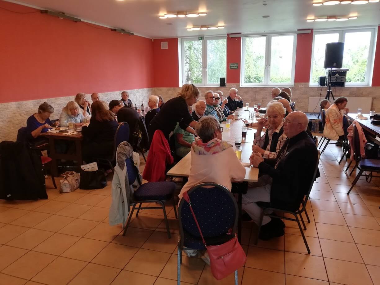 Ältere und jüngere Menschen sitzen in einem großen Raum mit orangen Wänden und mehreren Fenstern an langen Tischen. Sie essen, trinken und unterhalten sich. Auf einem Tisch stehen Getränke und Teller mit Speisen.