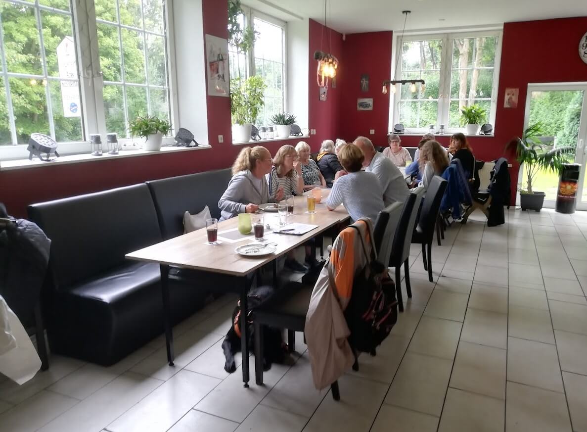 Mehrere Personen sitzen an zwei zusammengestellten Tischen in einem Restaurant mit roten Wänden und großen Fenstern. Sie unterhalten sich und trinken. Unter einem Stuhl liegt ein schwarzer Hund, vermutlich ein Blindenführhund.