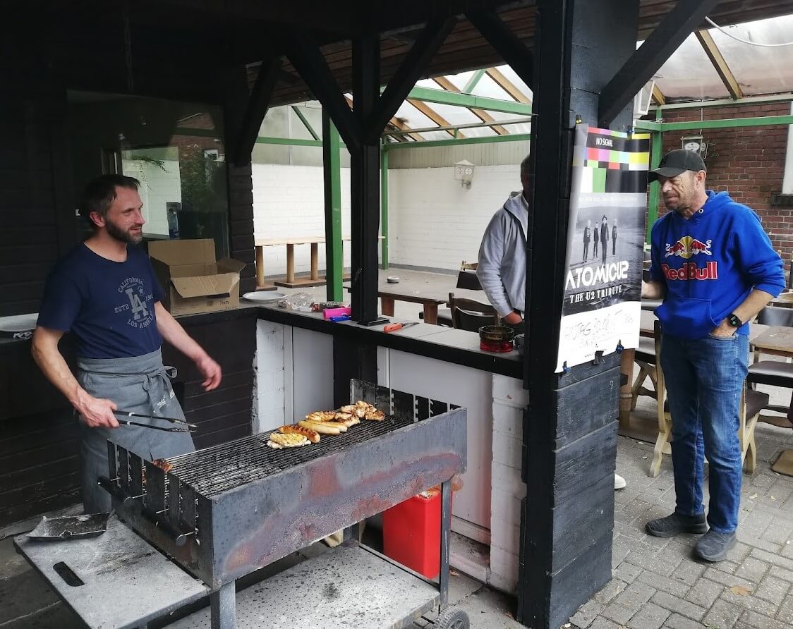 Ein Mann steht an einem großen Holzkohlegrill unter einem überdachten Außenbereich und grillt Würstchen und Fleisch. Neben ihm steht ein Mann in einem blauen Red-Bull-Hoodie, der ihm zusieht. Im Hintergrund ist ein teilweise überdachter Sitzbereich mit Holztischen und -stühlen zu sehen.