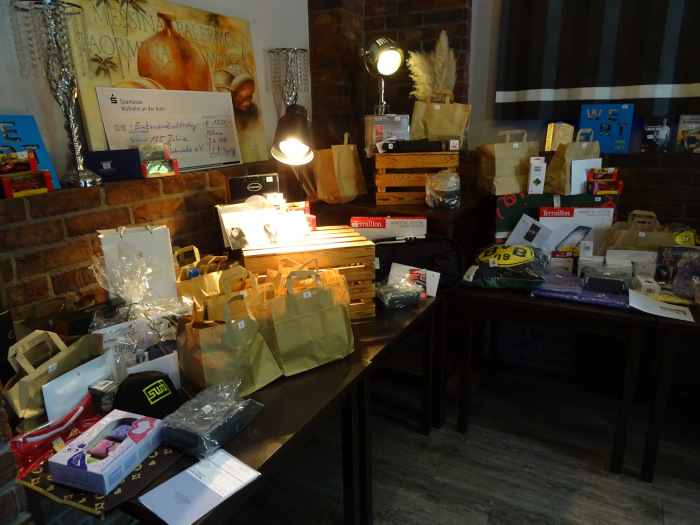 Auf mehreren Tischen sind zahlreiche Preise für eine Tombola ausgestellt. Zu sehen sind Geschenktüten, Gutscheine, Spiele, Fanartikel, Haushaltswaren und weitere Sachpreise. Im Hintergrund stehen ein großer symbolischer Spendenscheck sowie dekorative Leuchten und Holzkisten. Die Preise sind übersichtlich für die Teilnehmerinnen und Teilnehmer der Tombola präsentiert.