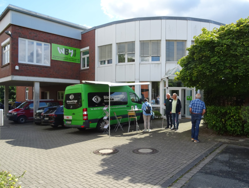 Vor dem Eingang eines modernen Gebäudes mit der Aufschrift „WBH – Westdeutsche Blindenhörbücherei“ steht ein grüner Kleinbus mit dem Schriftzug „Blickpunkt Auge“. Davor befinden sich ein Stehtisch und einige Informationsmaterialien. Mehrere Personen stehen im Eingangsbereich und unterhalten sich, teilweise mit Blindenstock.