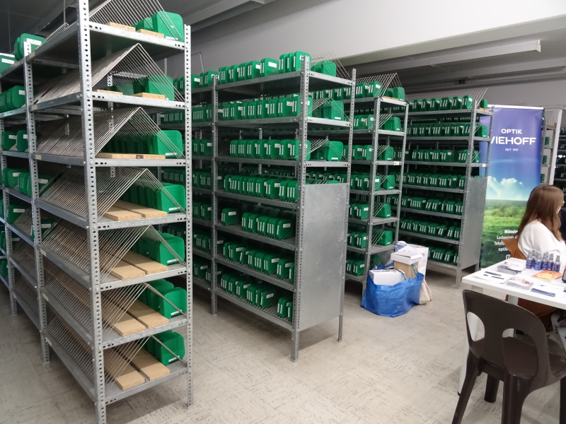 Ein Raum mit mehreren hohen Metallregalen, die grüne Kisten mit weißen Beschriftungen enthalten. Die Regale sind ordentlich sortiert und vermitteln den Eindruck eines umfangreichen Archivs oder Lagersystems. Rechts im Bild sitzt eine Frau an einem Infostand mit Werbematerial und einem Banner mit der Aufschrift „Optik Viehoff“.