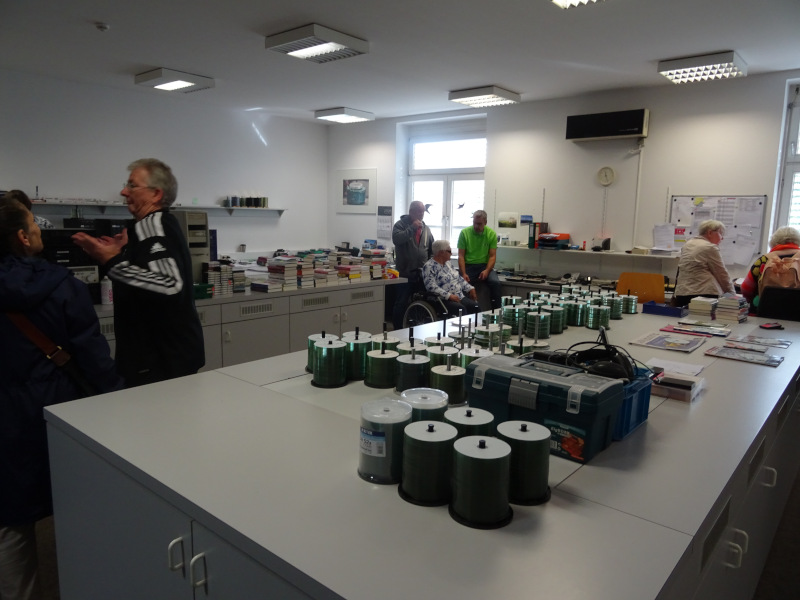 Ein heller Arbeitsraum mit weißen Wänden und mehreren Personen, die sich unterhalten oder arbeiten. Auf den Tischen liegen zahlreiche gestapelte CDs sowie Bücher. Im Hintergrund sitzt eine Frau im Rollstuhl, andere Besucher sehen sich im Raum um.