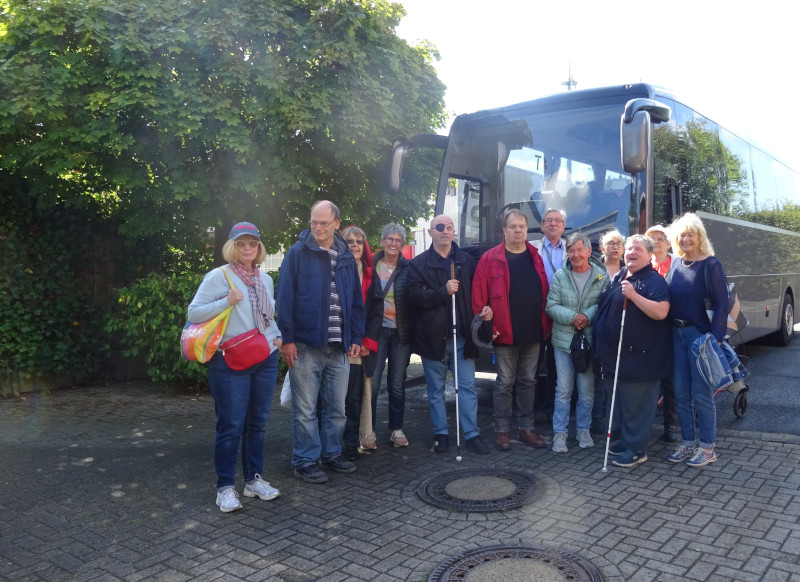 Eine Gruppe von zwölf Personen steht vor einem großen, modernen Reisebus auf einem Parkplatz. Einige der Personen haben Blindenstöcke, was darauf hinweist, dass sie sehbehindert sind. Im Hintergrund steht ein großer, dichter Baum, der Schatten spendet. Alle schauen in die Kamera und lächeln.