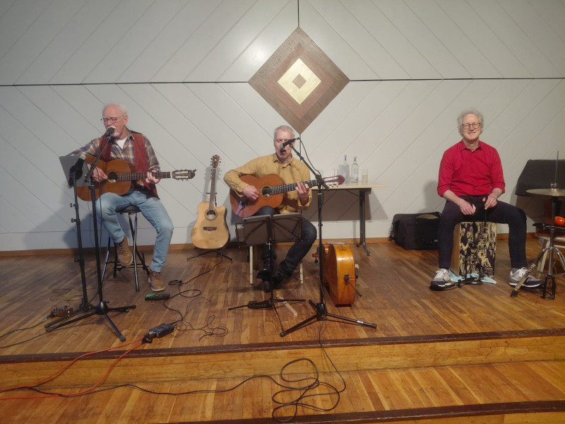 Drei ältere Männer sitzen auf einer Bühne und machen Livemusik. Zwei von ihnen spielen Gitarre und singen, der dritte sitzt auf einer Cajón und lächelt. Im Hintergrund stehen weitere Instrumente und Getränkeflaschen auf einem Tisch. Die Stimmung wirkt entspannt und musikalisch.