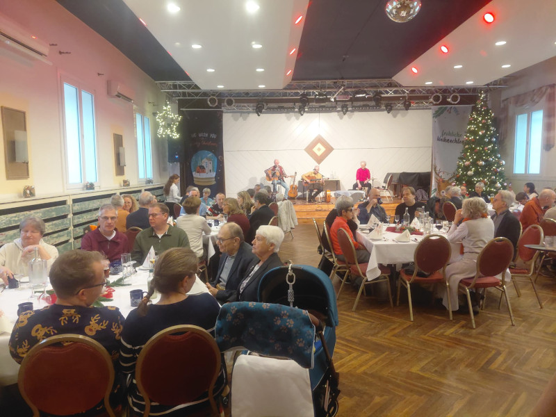 Ein großer Saal ist festlich für eine Veranstaltung geschmückt. An mehreren runden Tischen sitzen zahlreiche Gäste, darunter viele ältere Menschen, die sich unterhalten oder auf die Bühne schauen. Im Hintergrund spielen drei Musiker auf einer Bühne. Rechts steht ein geschmückter Weihnachtsbaum, was auf eine Weihnachtsfeier hinweist.