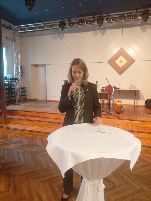 Die stellvertretende Bürgermeisterin Alexandra Hanf steht hinter einem runden Stehtisch mit weißer Tischdecke und spricht in ein Mikrofon. Sie trägt einen schwarzen Blazer über einer gemusterten Bluse. Im Hintergrund befindet sich eine kleine Bühne mit Musikinstrumenten und einem Tisch. Die Szene spielt sich in einem festlich eingerichteten Veranstaltungssaal ab.